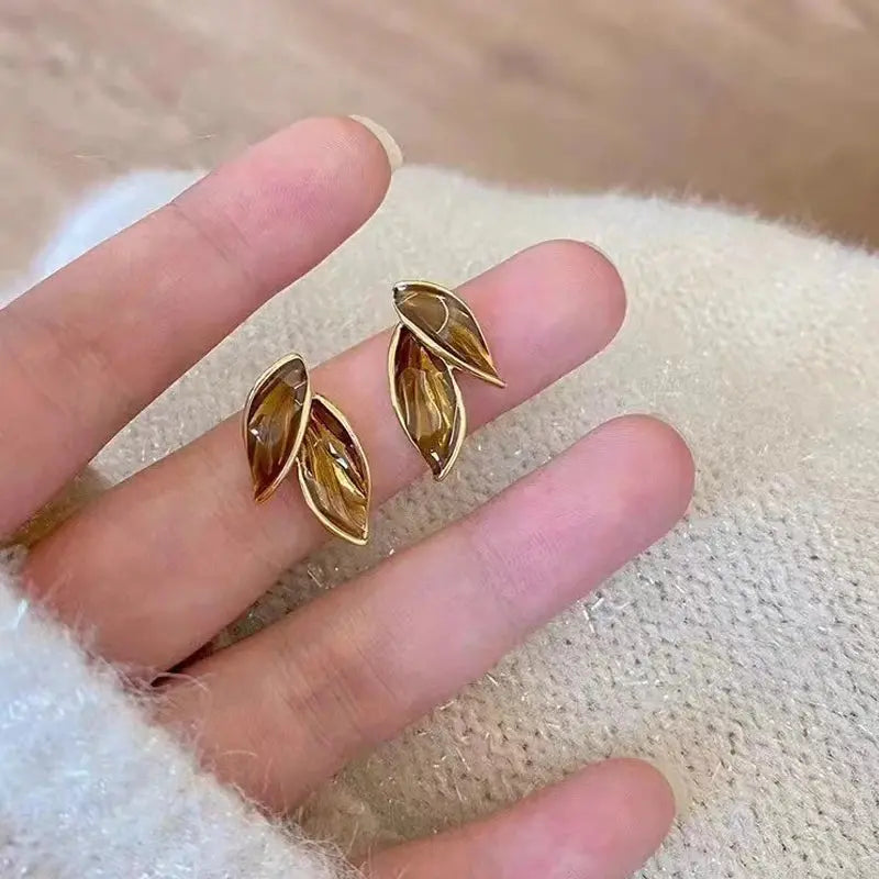 Gold leaf-shaped earrings on a finger against a textured beige background