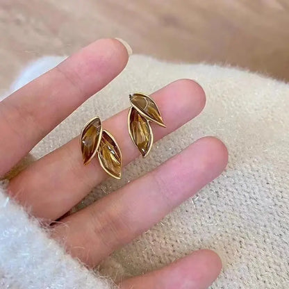 Gold leaf-shaped earrings on a finger against a textured beige background