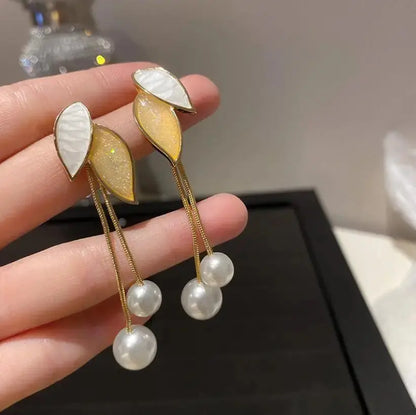 Earrings with leaf-shaped pendants and pearl drops held by a hand against a neutral background.